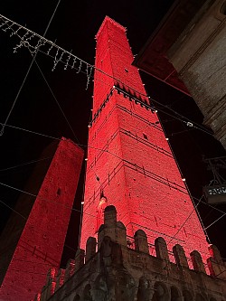 Asinelli and Garisenda Bologna towers. In the heart of Bologna, Italy, two defiant towers pierce the sky, casting long shadows in the golden afternoon light. The taller, Asinelli, stands proud, its ancient brickwork worn smooth by centuries. Garisenda, its companion, leans precariously to the side, a testament to both the passage of time and the forces of nature.   A bright yellow ribbon bisects Garisenda, a stark reminder of its precarious dance with gravity. It remains on 